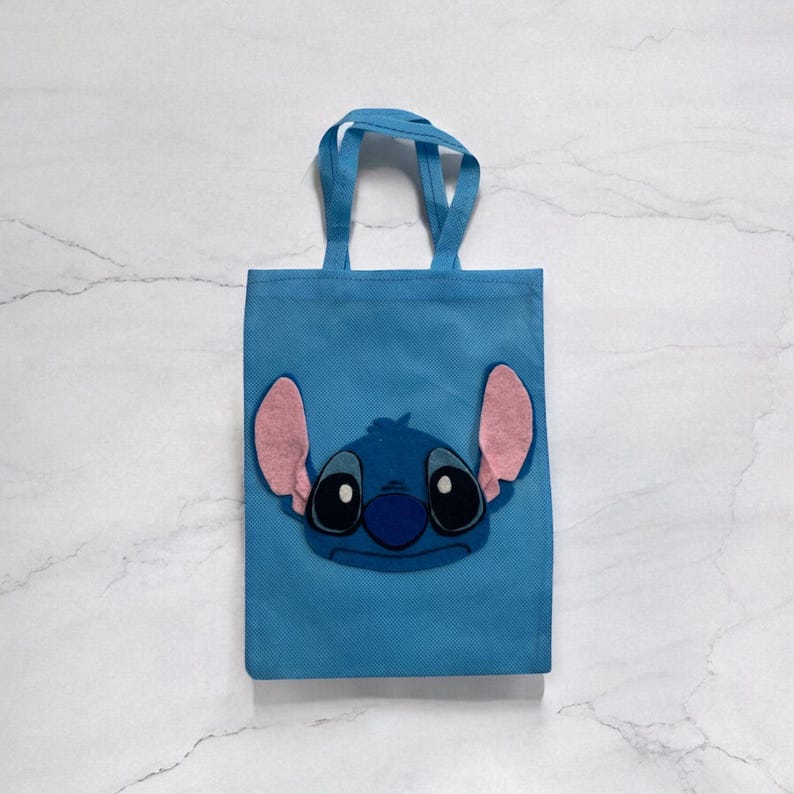 May include: A light blue tote bag with a cartoon character's face, large black eyes, a blue nose, and pink ears. The bag has matching blue handles and is made of a non-woven fabric. The character's face is a felt applique.