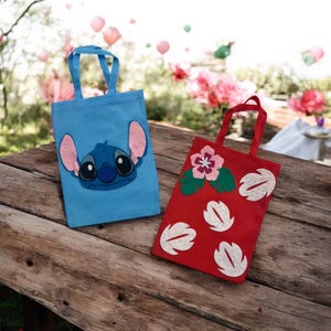 May include: Two tote bags on a wooden surface. One is light blue with a Stitch character design, the other is red with a floral and leaf design. Both bags have matching handles.
