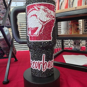 Puede incluir: Un vaso negro con asa, completamente cubierto de peque&ntilde;os diamantes de imitaci&oacute;n negros, rojos y blancos. El dise&ntilde;o presenta una silueta de jabal&iacute; blanca sobre fondo rojo y la palabra Razorbacks escrita en letra cursiva blanca en la parte inferior.