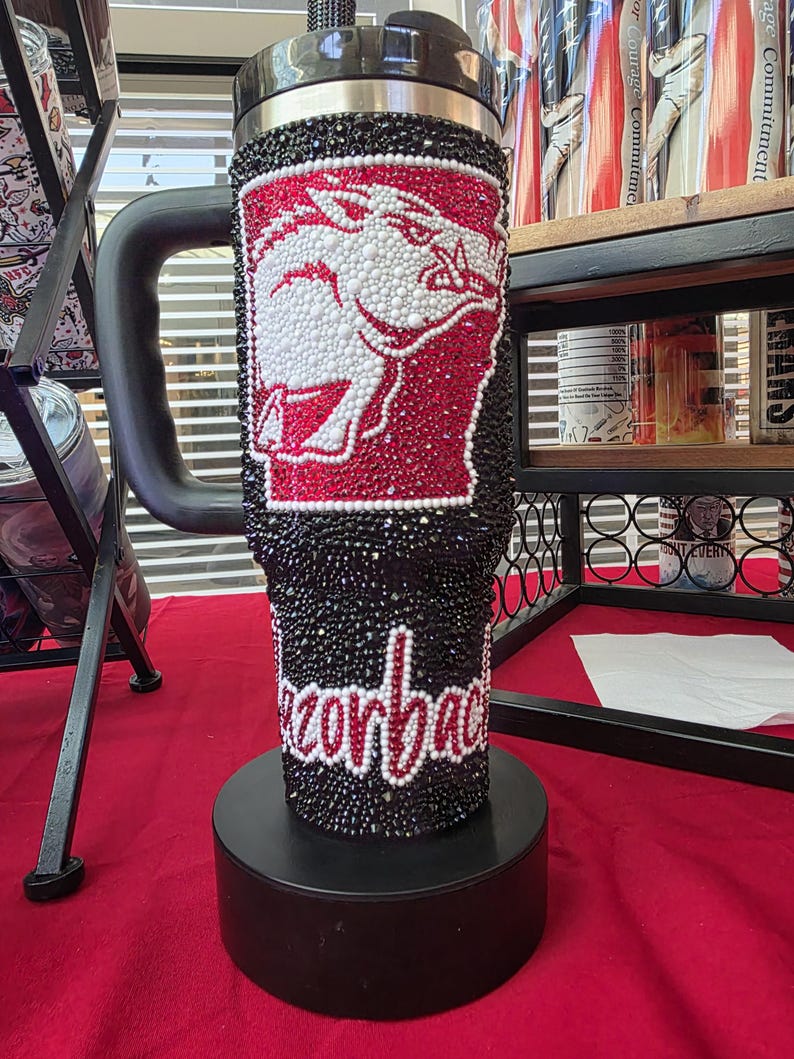 Puede incluir: Un vaso negro con asa, completamente cubierto de peque&ntilde;os diamantes de imitaci&oacute;n negros, rojos y blancos. El dise&ntilde;o presenta una silueta de jabal&iacute; blanca sobre fondo rojo y la palabra Razorbacks escrita en letra cursiva blanca en la parte inferior.