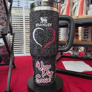 Puede incluir: Un vaso Stanley negro cubierto de peque&ntilde;os diamantes de imitaci&oacute;n negros con un dise&ntilde;o de coraz&oacute;n hecho de diamantes blancos y rojos. El texto "Woo Pig Sooie" est&aacute; escrito en diamantes rojos y blancos debajo del coraz&oacute;n.