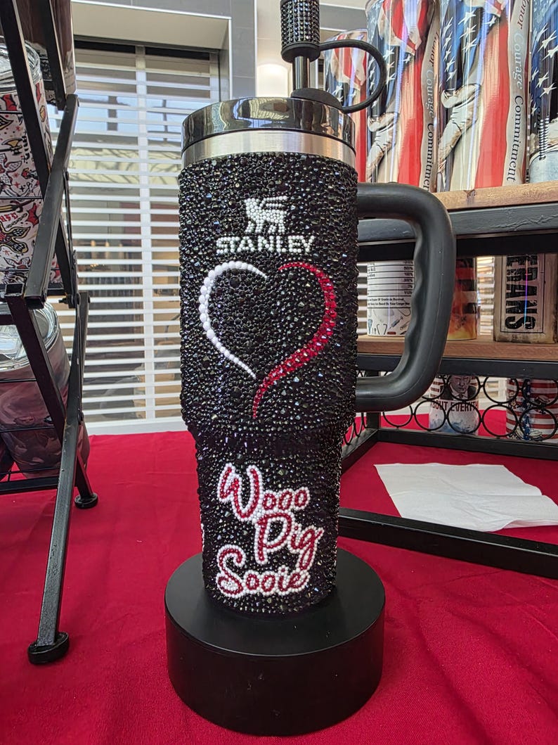 Puede incluir: Un vaso Stanley negro cubierto de peque&ntilde;os diamantes de imitaci&oacute;n negros con un dise&ntilde;o de coraz&oacute;n hecho de diamantes blancos y rojos. El texto "Woo Pig Sooie" est&aacute; escrito en diamantes rojos y blancos debajo del coraz&oacute;n.