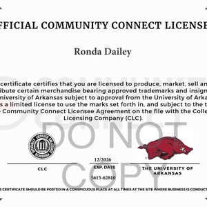 Puede incluir: Un certificado oficial de licenciatario de Community Connect para Ronda Dailey, que autoriza la producci&oacute;n de mercanc&iacute;a de la Universidad de Arkansas. El documento presenta el logotipo de razorback de la Universidad de Arkansas, un sello CLC y fecha de vencimiento 12/2026.