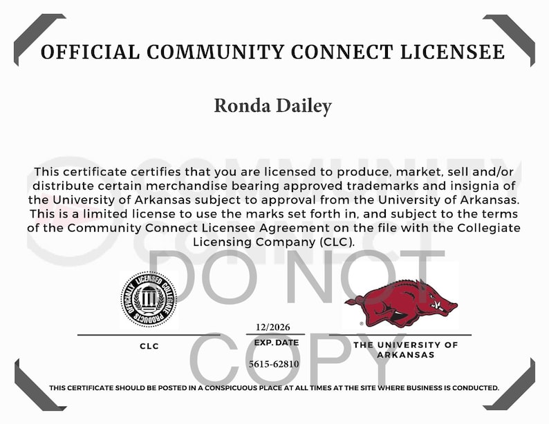 Puede incluir: Un certificado oficial de licenciatario de Community Connect para Ronda Dailey, que autoriza la producci&oacute;n de mercanc&iacute;a de la Universidad de Arkansas. El documento presenta el logotipo de razorback de la Universidad de Arkansas, un sello CLC y fecha de vencimiento 12/2026.