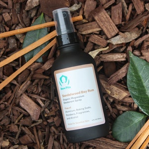 May include: A black bottle of Sandalwood Bay Rum organic magnesium deodorant spray. The bottle is labeled with the brand name "Bare Pits" and lists ingredients, including "Free of Aluminum, Baking Soda, Parabens, Fragrances, and Alcohol". The bottle is sitting on a bed of brown wood chips.