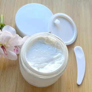 May include: A white jar of face cream with a white lid and a white spatula. The jar is open and the cream is visible.