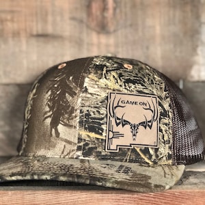 May include: A brown and green camouflage trucker hat with a brown mesh back. The hat has a brown leather patch with a white outline and the text "GAME ON" in white. The patch also features a stylized image of a deer skull with antlers.
