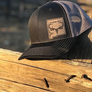 May include: A black and gray trucker hat with a brown leather patch that says "GAME ON" and features a stylized deer head and the outline of the state of New Mexico.