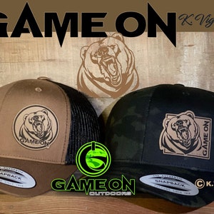 May include: Two brown and black trucker hats with a brown bear graphic and the text "GAME ON" on the front. The hats are on a wooden surface.
