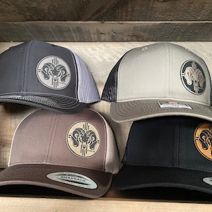 May include: Four different color baseball caps with a brown leather patch featuring a ram's head and a Native American symbol. The caps are black, brown, gray, and black with a brown bill.