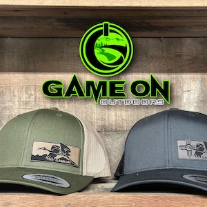 May include: Two trucker hats with the text "GAME ON OUTDOORS". One hat is olive green with a tan mesh back and a patch with a bird graphic. The other hat is charcoal gray with a black mesh back and a patch with a sun graphic.