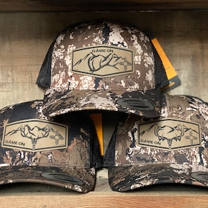 May include: Three camouflage trucker hats with a brown leather patch that says "GAME ON" and a graphic of a mountain range and an elk skull.