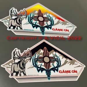 May include: Two white diamond-shaped stickers with a black and white elk, a turquoise and black skull with antlers, and a red and white sun symbol. The stickers have a black and white mountain range in the background and the text "GAME ON" in red and white. The stickers have a copyright notice at the bottom.