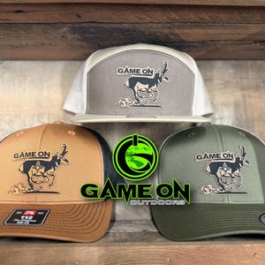 May include: Three baseball caps in tan, olive green, and light gray, each featuring a graphic of a running pronghorn and the text "GAME ON". The caps have mesh back panels and structured crowns. A bright green circular logo with the text "GAME ON OUTDOORS" is centered.