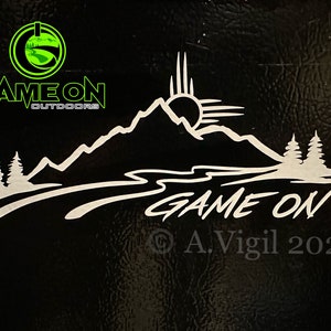 May include: White vinyl decal with a mountain range, a river, and two pine trees. The sun is rising over the mountains. The text "GAME ON" is written in a stylized font below the mountains. The text "A. Vigil 2023" is written in a smaller font below the text "GAME ON".