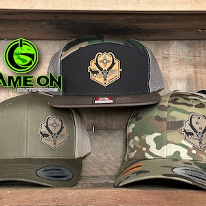 May include: Three camouflage trucker hats with a brown leather patch featuring a skull and antlers. The patch has the text "GAME ON" on it. The hats are on a wooden surface.