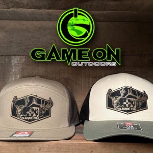 May include: Two baseball caps featuring a hexagonal patch with a deer graphic and the text GAME ON. The left cap is tan with a mesh back, and the right cap is cream with a black mesh back and olive brim. Above them is a neon green logo with GAME ON OUTDOORS.