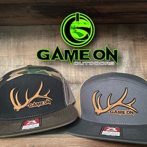 May include: Two black and brown baseball caps with a brown antler graphic and the text "GAME ON" on the front. The caps have a mesh back and a flat bill.