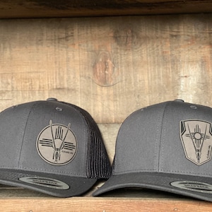 May include: Two black trucker hats with a black mesh back. The hats have a gray leather patch on the front with a stylized design of a sun, a symbol, and an animal head. The text "GAME ON" is on the patch.