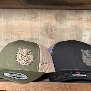 May include: Two trucker hats with a moose graphic and the text "Game On". One hat is green with a tan mesh back, and the other is black with a gray mesh back.