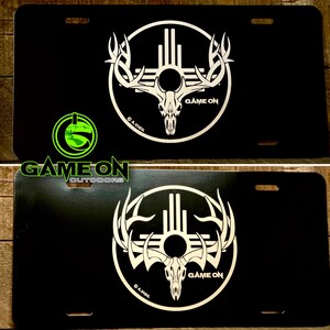May include: Black license plate featuring a white graphic of a deer skull with antlers inside a circle. The words "GAME ON" and "© A.VIGIL" are also visible. The logo "GAME ON OUTDOORS" is in the lower left corner.