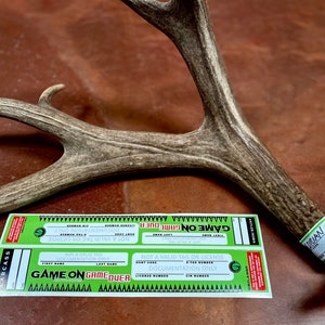 May include: A deer antler with attached green and white tags. The tags read "GAME ON GAME OVER" and include fields for documentation. The antler is a natural brown color and rests on a brown surface.