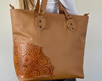 May include: A tan leather tote bag with a floral design embossed on the front. The bag has two top handles and a zipper closure.