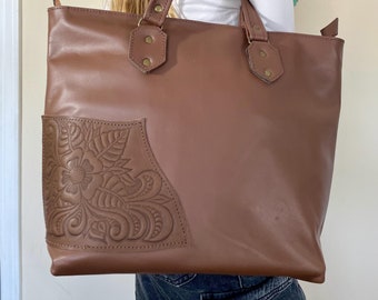 May include: A brown leather tote bag with a floral design embossed on the front pocket. The bag has two top handles and a zipper closure.