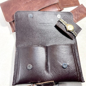 K&ouml;nnte beinhalten: Ein braunes Lederuhrenetui mit zwei F&auml;chern und einem Druckknopfverschluss. Das Etui tr&auml;gt ein Etikett mit der Aufschrift "Collections by".