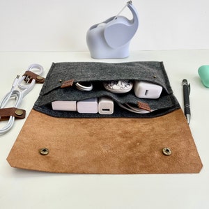 May include: A gray felt pouch with a brown leather flap and snaps. The pouch is open and filled with various electronic cords and chargers.