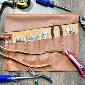 May include: A brown leather tool roll containing a selection of tools, including wrenches, pliers, a utility knife, a screwdriver, and a tape measure. The roll is unfurled, revealing the tools within.