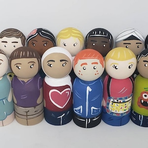 May include: A set of 12 wooden peg dolls, each painted with a different face and outfit. The dolls are all wearing different clothes, including a blue jacket, a red shirt, a purple shirt, and a green shirt. The dolls are all smiling and have different hair colours.