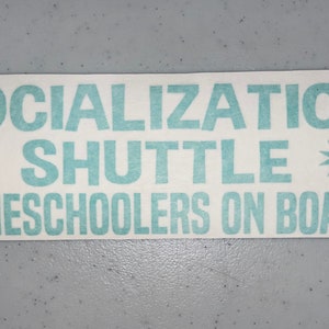 May include: A rectangular white vinyl decal featuring teal lettering that reads SOCIALIZATION SHUTTLE and HOMESCHOOLERS ON BOARD, with a teal starburst symbol on each side of the word SHUTTLE, designed for application on a smooth surface.