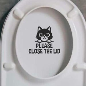 Può includere: Un sedile del water bianco con un adesivo bianco e nero che dice "Per favore, chiudi il coperchio". Un gatto nero dei cartoni animati con occhi bianchi sta guardando da sopra l'adesivo.
