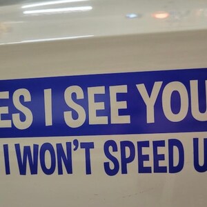 May include: A rectangular white bumper sticker featuring a bold blue horizontal stripe. The text is printed in white capital letters reading YES I SEE YOU! on the top line and NO I WON'T SPEED UP! in blue letters below.