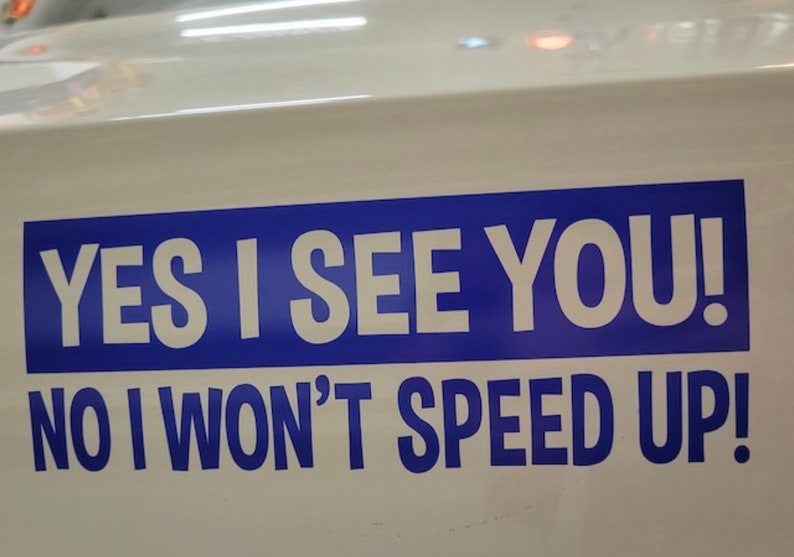 May include: A rectangular white bumper sticker featuring a bold blue horizontal stripe. The text is printed in white capital letters reading YES I SEE YOU! on the top line and NO I WON'T SPEED UP! in blue letters below.
