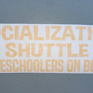 May include: A rectangular adhesive decal featuring the light orange text SOCIALIZATION SHUTTLE HOMESCHOOLERS ON BOARD in a bold, sans-serif font. The design includes a small, eight-pointed star icon on both the left and right sides of the word SHUTTLE.