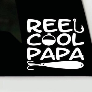 May include: White vinyl decal featuring the text "REEL COOL PAPA" in a bold font. The design includes a fishing hook, a bobber, and a lure, all in white, set against a black background.