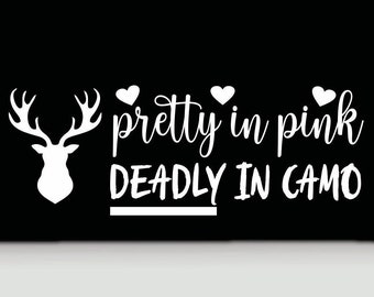 Puede incluir: Una calcomanía negra con una silueta blanca de una cabeza de ciervo con astas a la izquierda. El texto dice "pretty in pink" en fuente cursiva con tres corazones pequeños encima, y "DEADLY IN CAMO" en fuente negrita sin remates debajo.