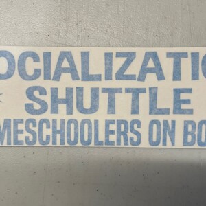 May include: A rectangular white vinyl sticker featuring light blue text that reads SOCIALIZATION SHUTTLE HOMESCHOOLERS ON BOARD. The design includes two light blue star-shaped icons positioned on either side of the word SHUTTLE.
