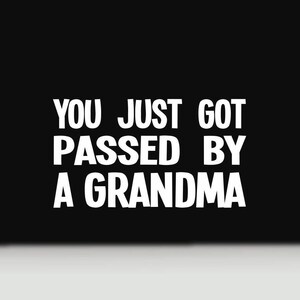 May include: A black rectangular decal featuring the white text YOU JUST GOT PASSED BY A GRANDMA in a bold, sans-serif font. The lettering is arranged in four lines, centered against the solid black background, creating a high-contrast graphic design.
