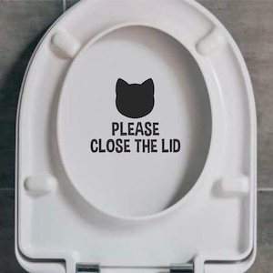 Può includere: Un sedile del water bianco con una silhouette di gatto nero e il testo "Si prega di chiudere il coperchio".