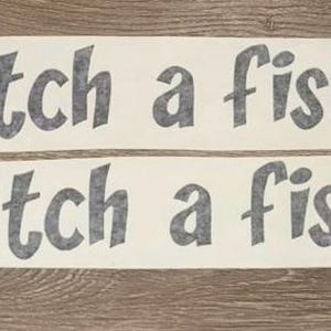 May include: Two rectangular adhesive decals featuring the dark grey text Catch a fishy in a bold, rounded sans-serif font. The lettering is printed on a white background, presented as two separate horizontal strips for application.