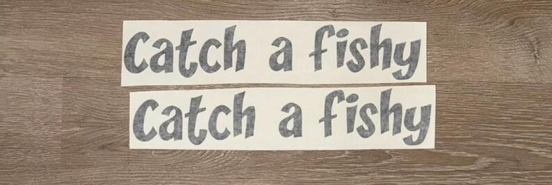 May include: Two rectangular adhesive decals featuring the dark grey text Catch a fishy in a bold, rounded sans-serif font. The lettering is printed on a white background, presented as two separate horizontal strips for application.