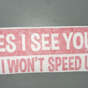 May include: A rectangular bumper sticker with a red background and white text that reads YES I SEE YOU! NO I WON'T SPEED UP! The lettering is in a bold, sans-serif font, and the sticker has a clean, white border around the red rectangular area.