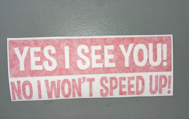 May include: A rectangular bumper sticker with a red background and white text that reads YES I SEE YOU! NO I WON'T SPEED UP! The lettering is in a bold, sans-serif font, and the sticker has a clean, white border around the red rectangular area.
