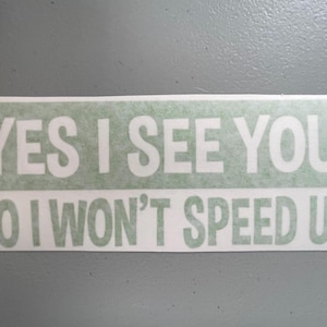 May include: A rectangular bumper sticker with a light green background and white text that reads YES I SEE YOU! NO I WON'T SPEED UP! The sticker features bold, sans-serif lettering arranged in two lines with an exclamation point at the end.