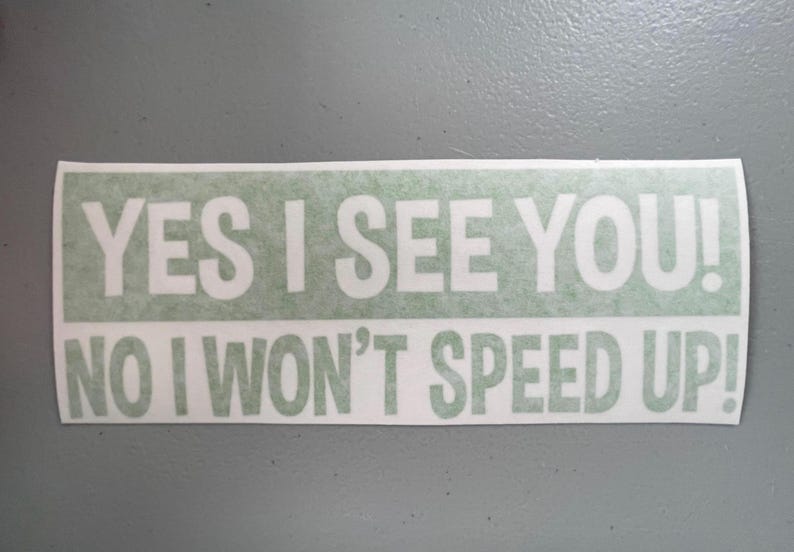 May include: A rectangular bumper sticker with a light green background and white text that reads YES I SEE YOU! NO I WON'T SPEED UP! The sticker features bold, sans-serif lettering arranged in two lines with an exclamation point at the end.