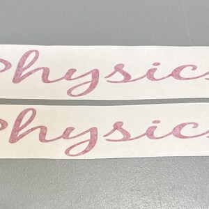 May include: Two identical white rectangular adhesive decals with pointed ends, each featuring the word Physics printed in a cursive, dusty rose-coloured font. The lettering has a textured, slightly mottled appearance across the surface.