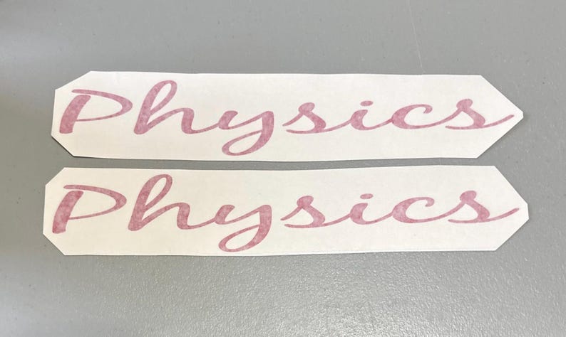 May include: Two identical white rectangular adhesive decals with pointed ends, each featuring the word Physics printed in a cursive, dusty rose-coloured font. The lettering has a textured, slightly mottled appearance across the surface.